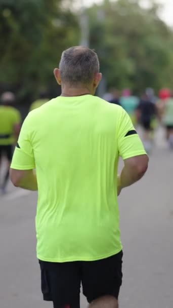 縦画面 市内マラソンを実行している黄色のTシャツを着て灰色の髪を持つスローモーションのシニア男 彼の前に他の出場者 スポーツイベントに参加する競技者を撮影した後 — ストック動画