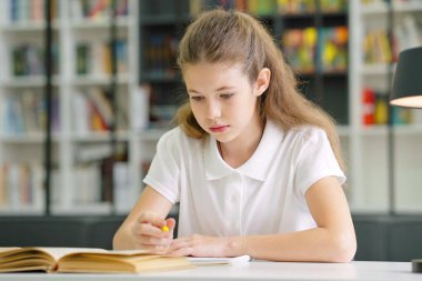 Intelligent school girl sitting at desk and writing in exercise book, lamp beside her, bookshelves on blurred background. Pupil doing homework. Concept of education