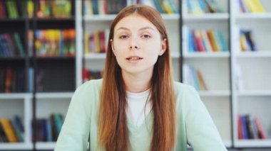 El sallayan genç kız kütüphanede kameraya bakarak konuşuyor. Kafkasyalı ergen vlog 'u kaydediyor, sosyal medya için yayın yapıyor, online sohbet için video arıyor. Web kamerası görüntüsü. Uzaklık öğrenimi