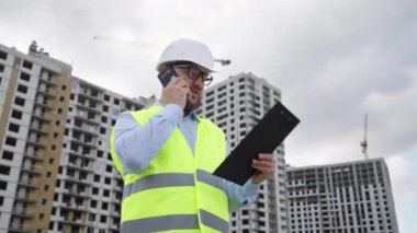 Sakallı kendine güvenen mühendis sabit şapka ve güvenlik yeleği giyiyor inşaat alanında cep telefonuyla konuşuyor, önemli konuları tartışıyor, arka planda çok katlı binalar ve vinçler var.
