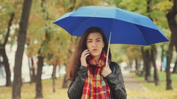 雨の中 傘の下で秋の公園に立って電話で話している小切手のスカーフを身に着けている若い女性 背景にぼやけた木 — ストック動画
