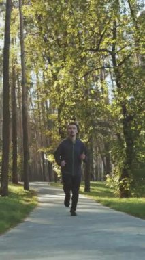 Kıvırcık saçlı, spor ceketli, parkta yeşil ağaçların arasında yolda koşan yavaş çekim sporcusu. Atış talimi sabah güneşinde. Zindelik kavramı