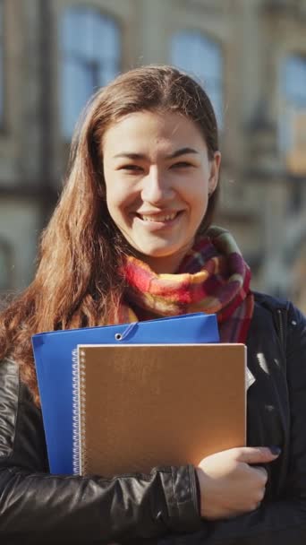 垂直屏风 快乐的女人 在阳光灿烂的日子穿着皮夹克和围巾 手里拿着书本 模糊的大学建筑背景 女学生的肖像 教育的概念 — 图库视频影像