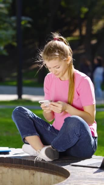 縦型スクリーン カジュアルな服を着た若い女性がベンチに座って スマートフォン ノートパソコンの横に立って 白い鳩は彼女の隣に着陸する上でのテキストメッセージを足 学生と好奇心旺盛な鳥 — ストック動画