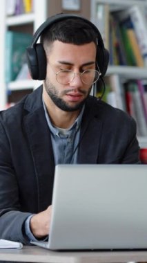 Dikey Ekran: Gözlüklü sakallı adam video görüşmesi için kulaklık ve laptop kullanıyor, webcam ile konuşuyor, telekonferans yapıyor veya kütüphanede yakınlaştırma yapıyor. Uzaklık öğrenimi