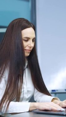 Vertical Screen: Female with long hair wearing white shirt working on laptop in office. Business woman reacting emotionally after receiving great news. Concept of success