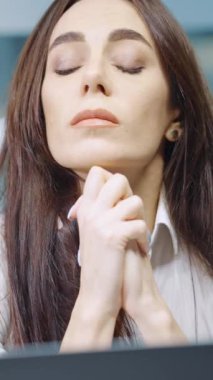 Vertical Screen: Sad woman with long hair wearing white shirt sitting in office in front of laptop, frowning and putting head on hands. Employee having problem at work. Concept of stress