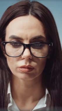 Vertical Screen: Pensive woman with long hair wearing eyeglasses working in office. Closeup portrait of businesswoman winking while reading documents or typing on laptop. Concept of occupation