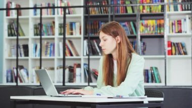 Young attractive university student studying at library. Caucasian teenager writing notes using laptop, planning working process. Exam preparation