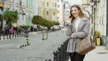 Cheerful blonde woman wearing casual gray coat talking on phone, blurred old building background. Young female waiting for taxi in city. Concept of communication