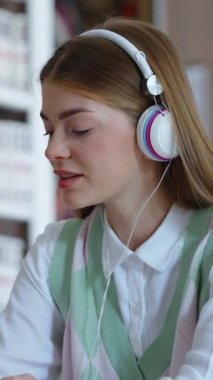 Vertical Screen: Cheerful student working on laptop in library and listening to favorite music with headphones, moving to the rhythm. Arc shot young female studying. Concept of education