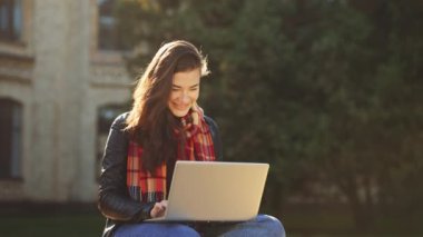 Deri ceket giyen, eşarplı ve kot pantolonlu genç bir kadın güneşli parkta oturuyor ve dizüstü bilgisayarla çalışıyor, bina geçmişini bulanıklaştırıyor. Uzak çalışma kavramı