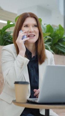 Vertical Screen: Cheerful employee sitting in cafe with laptop and talking on phone, coffee cup standing beside, businesswoman at coffee break, low angle tracking shot