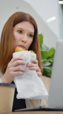 Vertical Screen: Businesswoman sitting in cafe, eating sandwich and watching video on laptop, employee having work break, low angle. Concept of food