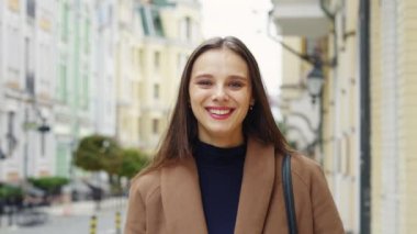 Closeup young happy wearing brown coat woman looking at camera in city. Caucasian female smiling charmingly. Beauty and femininity concept