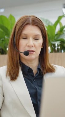 Vertical Screen: Adult woman online consultant using headset and laptop for communicating, business coach sitting in open space and performing training, tracking shot