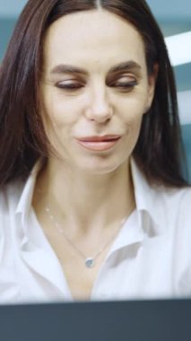 Vertical Screen: Female with long hair wearing white shirt looking at laptop screen and feeling joy while receiving great news, screaming and gesturing. Businesswoman in office. Concept of success