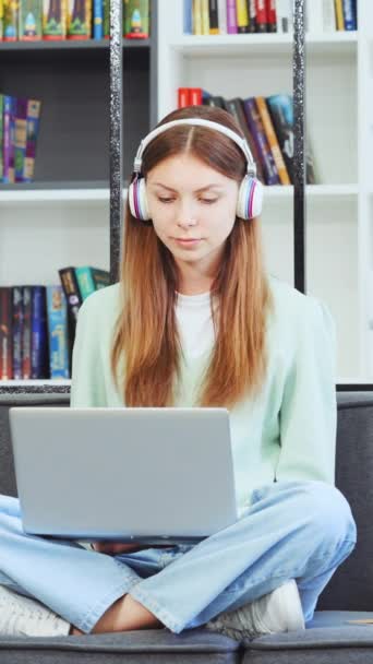 縦画面 キャンパス図書館でヘッドフォンで音楽を楽しむ女子大生 10代の女の子はノートパソコンと日の夢の割り当てを行う 喜び若いです女性歌彼女のお気に入りの歌 — ストック動画