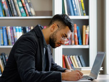 Sakallı erkek kütüphanede dizüstü bilgisayar kullanıyor, not alıyor, arka planda bulanık kitaplıklar var. Öğrenci ödevini hazırlıyor. Teknoloji ve eğitim kavramı