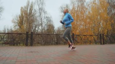 Fit blonde woman wearing blue sports jacket runnning jogging in autumn park. Tracking shot active female doing cardio training in the morning. Concept of healthy lifestyle