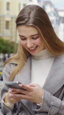 Vertical Screen: Smiling young woman writing sms online with smartphone. Dreamy stylish woman chatting at street, blurred old buildings on background. Mobile addiction concept