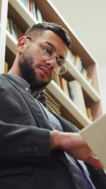 Dikey Ekran: Gözlüklü genç sakallı adam kitaplıkların yanında duruyor ve kitap okuyor, sayfaları çeviriyor. Kitapçıda veya kütüphanede edebiyattan keyif alan zeki bir okuyucunun düşük açılı portresi