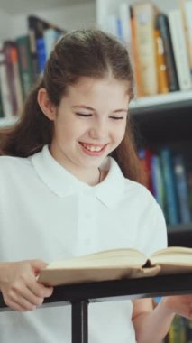 Vertical Screen: Child wearing white t-shirt standing by metal railings and reading book, stops reading and looking at camera smiling. Portrait of school girl in school library. Concept of education