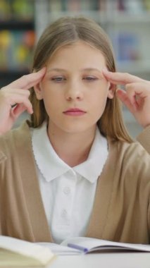 Vertical Screen: Sad school girl in casual wear sitting at table and suffering headache, massaging her head. Pupil feeling pains, symptom of disease. Concept of healthcare