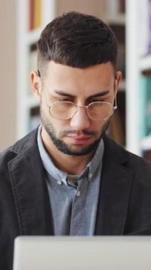 Vertical Screen: Young man in eyeglasses working on laptop in library, browsing internet, doing research, working remotely, studying online, bookcases on blurred background. Concept of education