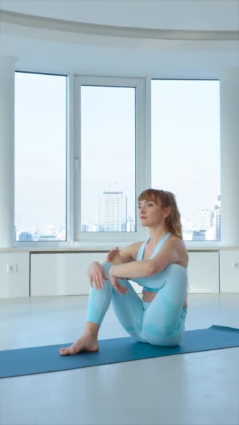 縦画面 ヨガスタジオで八角ポーズを練習している青いスポーツウェアの若いブロンドの女性 ヨギ女性アスタヴァクラサナに座って 腕の練習を行う上でバランスをとる 健康的なライフスタイルのコンセプト — ストック動画