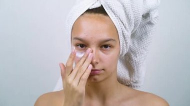 Adolescent girl with bath towel on head looking at camera and applying face cream on cheek. Time lapse pensive teenager with acne problems taking care of her skin. Concept of beauty