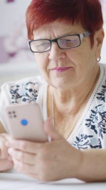Vertical Screen: Senior woman wearing eyeglasses sitting at table and using smartphone. Smiling aged person browsing internet or swiping photos in dating app. Concept of gadget