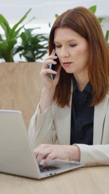 Vertical Screen: Businesswoman talking on phone and using laptop in open work space, green plants on wall behind her, remote worker in cafe, freelancer