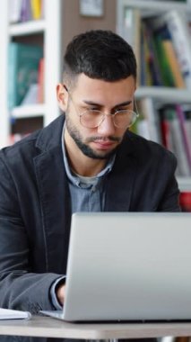 Vertical Screen: Handsome young man with eyeglasses sitting in library and browsing internet on laptop, looking stressed and unhappy. Distance learning. Remote work. Concept of technology