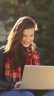 Deri ceket giyen, eşarp ve kot pantolon giyen, güneşli parkta oturan ve dizüstü bilgisayarla çalışan, binası bulanık genç bir kadın. Uzak çalışma kavramı