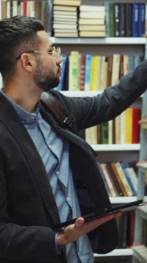 Dikey Ekran: Sakallı ve gözlüklü bir adam gömlek ve ceket giyiyor kitaplıkların yanından geçiyor ve elinde edebiyat listesi olan kitapları seçiyor. Kütüphanede üniversite öğrencisi