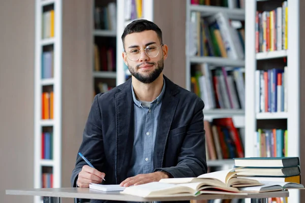 Young bearded male student with eyeglasses wearing jacket sitting in library and making notes on paper, looking up at camera and smiling. Portrait of intelligent man studying. Concept of education
