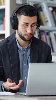 Dikey Ekran: Gözlüklü neşeli adam video görüşmesi için dizüstü bilgisayar ve kulaklık kullanıyor, webcam ile konuşuyor, el kol hareketi yapıyor, telekonferans yapıyor veya yakınlaştırma yapıyor, kütüphanede online eğitim alıyor.