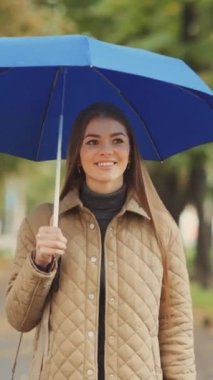 Mavi şemsiyeli, yağmurda gülümseyen genç bir kadın, şehir parkında yürüyor. Neşeli, güzel bir kadın yağmurlu havadan ve nemli iklimden hoşlanıyor. Hava kavramı