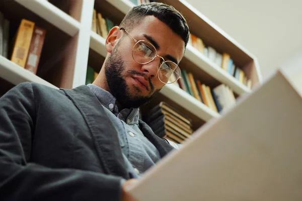 stock image Young bearded man with eyeglasses reading book while standing by bookcase in library, flipping pages, enjoying literature. Low angle reader going through interesting plot. Concept of education