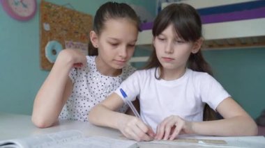 İki beyaz kız ödev yapıyor, dersleri tartışıyorlar. Öğrenciler evde ders çalışıyor. Bir kız diğerine yardım ediyor.