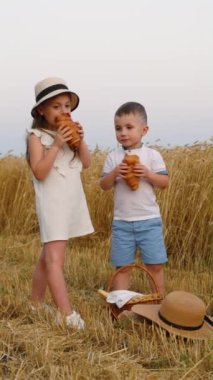 Yazlık giysiler giyen küçük çocuklar buğday tarlasının yanında durup kruvasan, sepet ve hasır şapka yiyorlar. Mutlu kardeşler hasat mevsiminde kırsalda yemek yiyor.