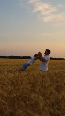 Yavaş çekim baba küçük oğlunu elleriyle tutuyor ve gün batımında buğday tarlasında dönüyor. Ark mutlu ebeveyn ve çocuğu birlikte eğlenirken çekti. Aile kavramı