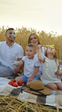 Dikey Ekran: Hasat mevsiminde buğday tarlasında sabun köpükleriyle piknik. Küçük çocuklar köpük üflüyor ve ebeveynler onları izliyor. Mutlu aile birlikte vakit geçiriyor. Eğlence anlayışı