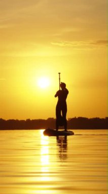 Dikey ekran: kadın silueti SUP tahtasının üzerinde dengede duruyor ve gün batımında kürek çekiyor, suya yansıyor. Açık hava aktivitesi ve sağlıklı yaşam tarzı. Zindelik kavramı