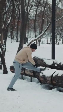 Kış parkında kar kaplı, spor kıyafetli, kütük üzerinde bacak esneten zinde bir adam. Dışarıda egzersiz yapan sporcuların arkası. Spor anlayışı