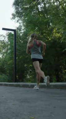 Dikey ekran: Ağır çekimde koşan kadın üstte ve parktaki yol kıvrımında koşan şortlar, yeşil ağaçların arasından parlayan güneş. Aktif bir kadın sabahları egzersiz yapıyor. Sağlıklı yaşam tarzı kavramı