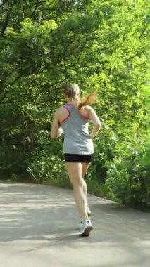 Dikey ekran: formalı kadın spor giysisi giyiyor dışarıda ağır çekimde koşuyor, asfalt yolun kenarında yeşil bitkiler ve ağaçlar var. Atış talimlerini takip ediyorum. Zindelik kavramı