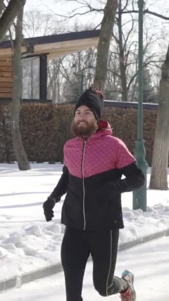 垂直屏风 男性慢跑者身穿粉色和黑色运动服 缓慢地在冬季公园里奔跑 地面上覆盖着雪 跟踪射击适合喜欢户外活动的人 体育的概念 — 图库视频影像