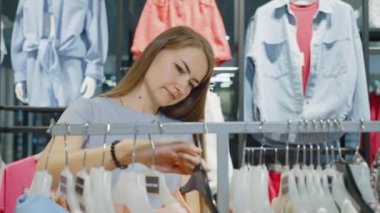 Moda mağazasında uzun saçlı, elbise askılarına dokunan beyaz bir kadın stil seçiyor ve kumaşı inceliyor. Uygun kıyafet arayan seçici bir müşteri. Alışveriş kavramı